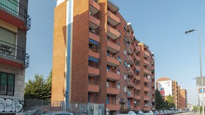 Un edificio residencial de varios pisos con balcones, un escape de incendios visible, y una vista de la calle con coches aparcados y cielo despejado.