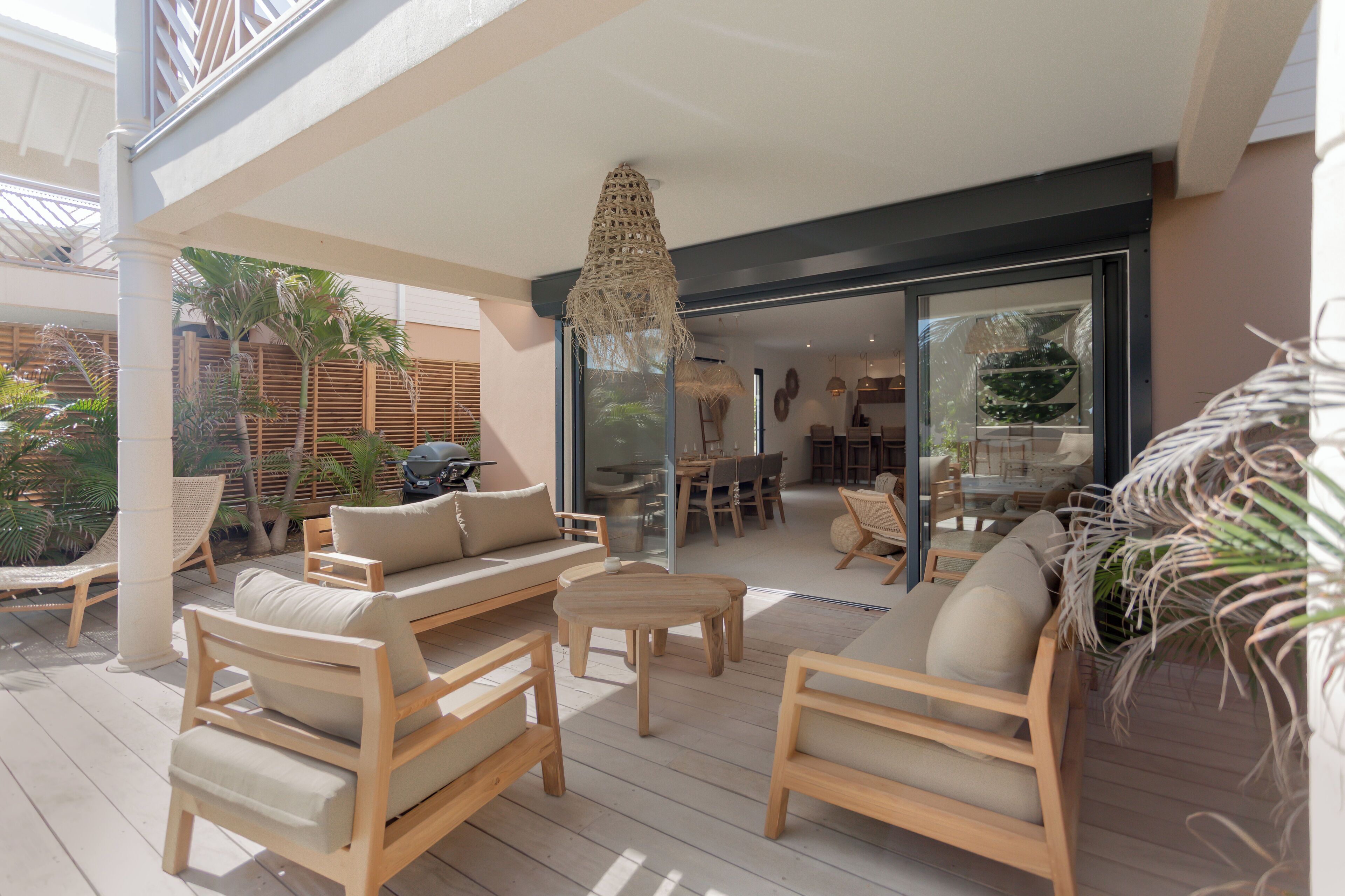 An outdoor patio area with wooden furniture, a woven ceiling light, and a view into the adjacent dining area.