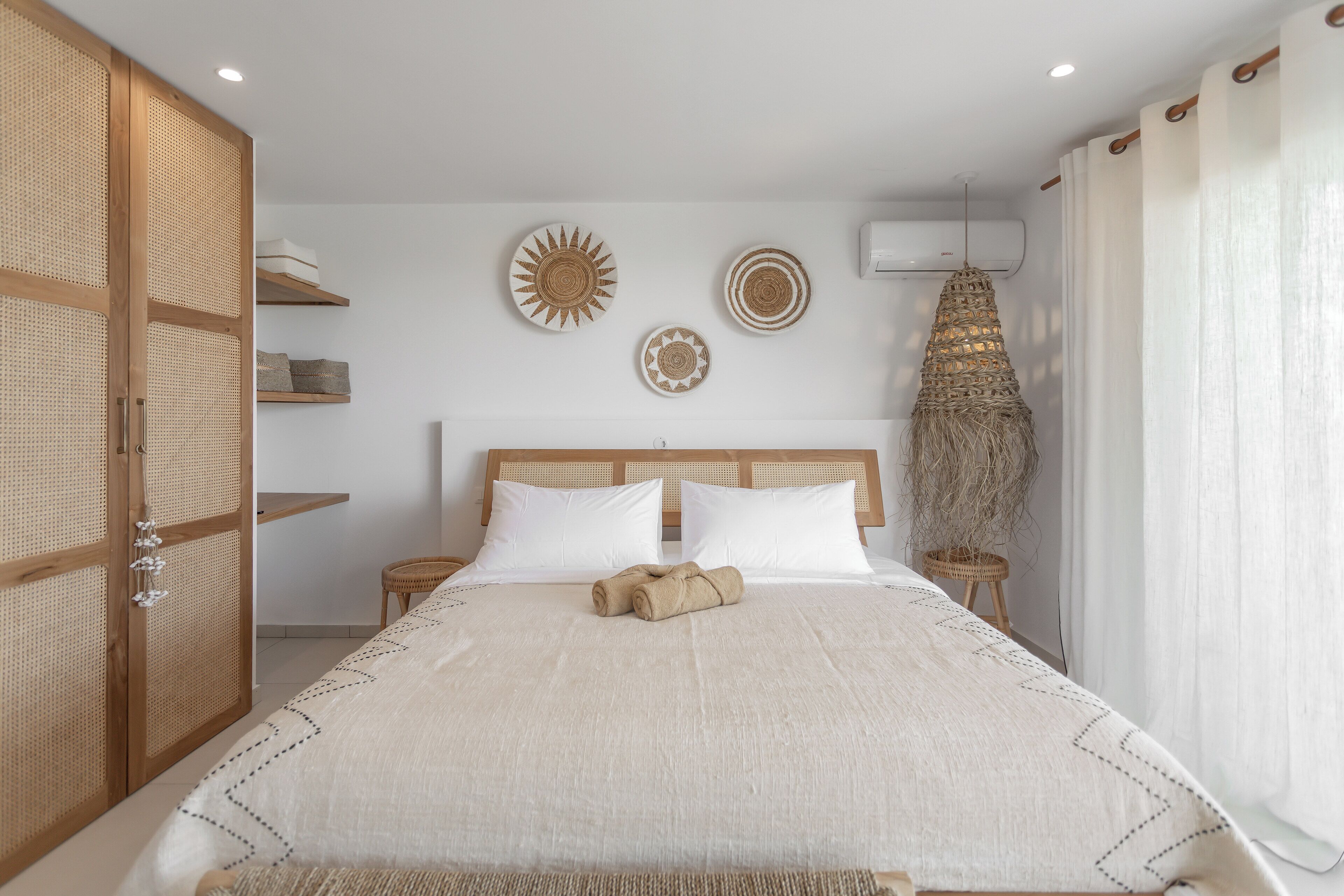 Un dormitorio ordenado con una cama grande, dos mesitas de noche, aire acondicionado de pared y una lámpara de fibras naturales.