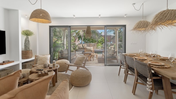 A modern dining area with a wooden table set for six, surrounded by wicker chairs and a large window offering a view of the outdoors. - Villa Aroa (Collectivité de Saint-Martin)