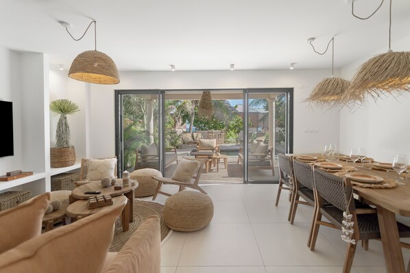 A modern dining area with a wooden table set for six, surrounded by wicker chairs and a large window offering a view of the outdoors.