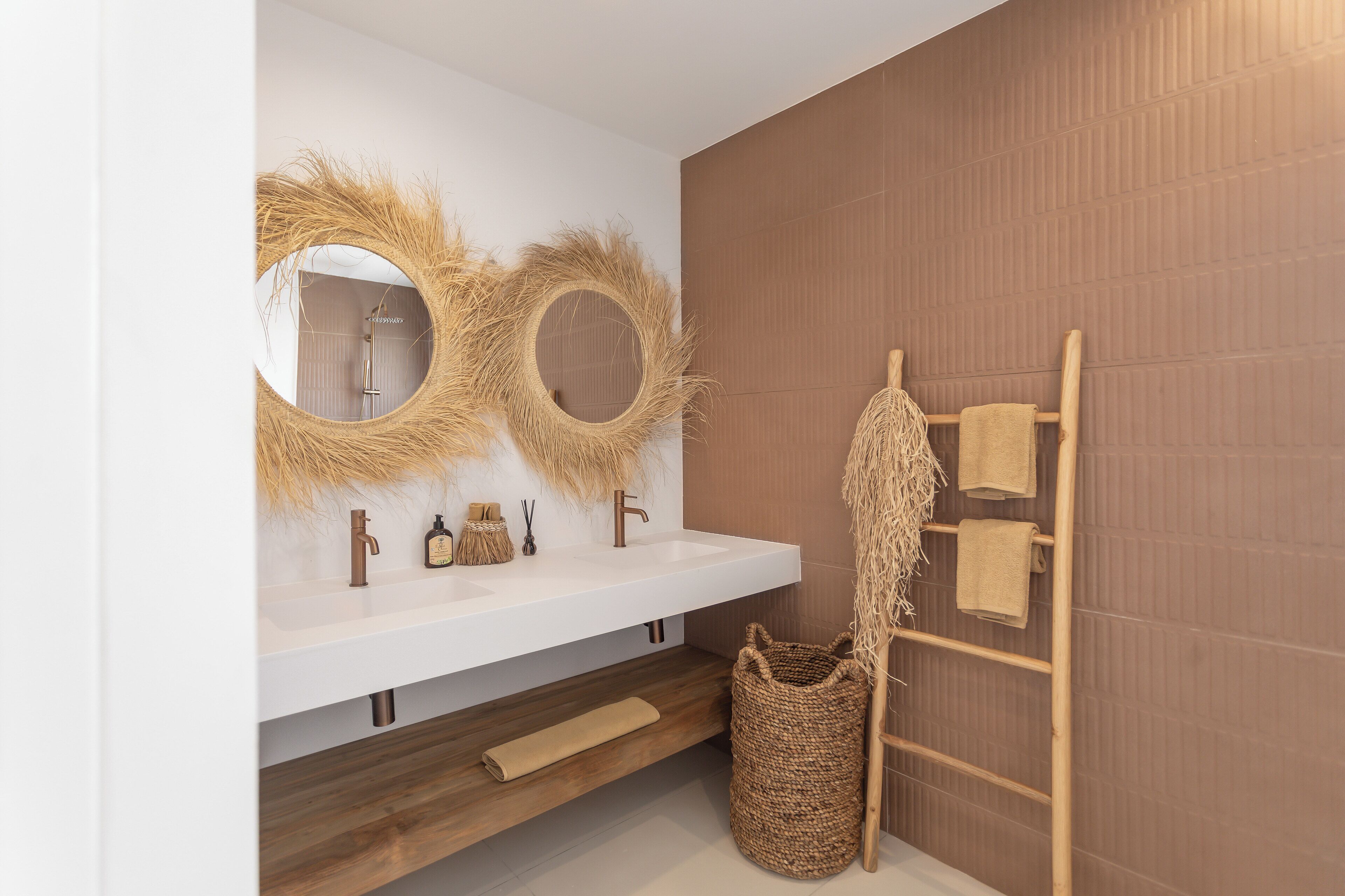 A modern bathroom with a white vanity, two round mirrors with natural fiber decorations, a wooden towel rack, and a woven basket.