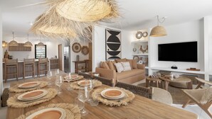 A modern living room with a dining area, featuring a large wooden table set for a meal, a sofa, and a television. - Villa Aroa (Collectivité de Saint-Martin)