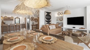 A modern living room with a dining area, featuring a large wooden table set for a meal, a sofa, and a television.