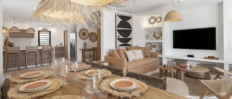 A modern living room with a dining area, featuring a large wooden table set for a meal, a sofa, and a television.