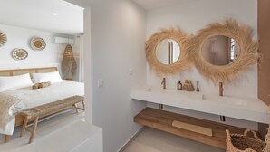 A modern bedroom with a large bed, a bedside bench, a wooden nightstand, and a bathroom with a white sink and two round mirrors. - Villa Aroa (Collectivité de Saint-Martin)