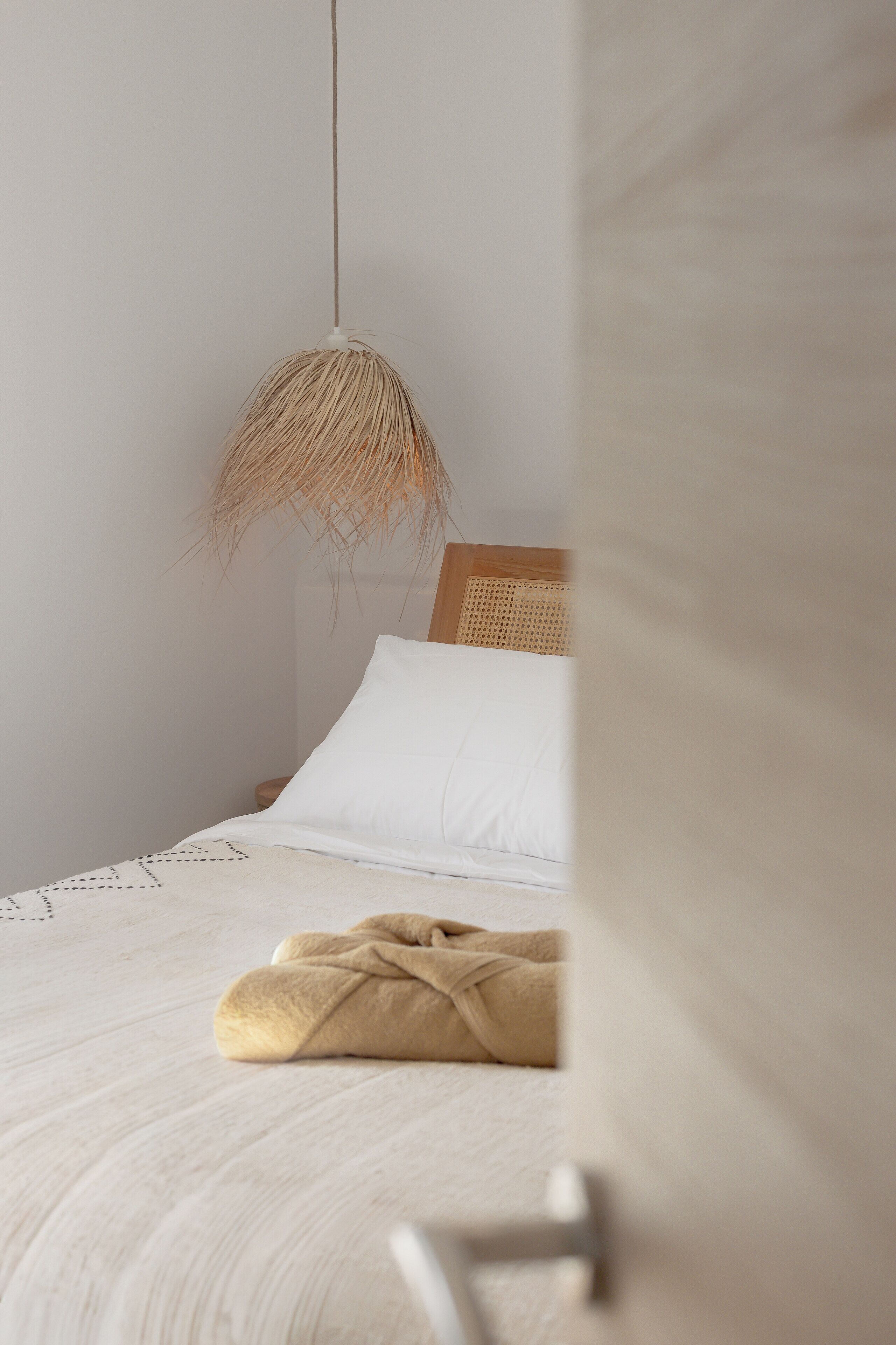 A neatly made bed with a white pillow and a beige blanket, a hanging light fixture, and a wooden headboard.