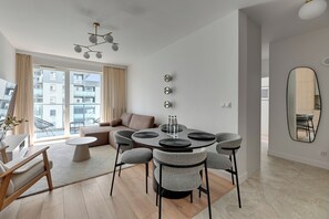 A modern living room with a round dining table, grey upholstered chairs, a sofa, and a large window with curtains. - Horizon 63 Superior by Grand Apartments (Gdańsk)