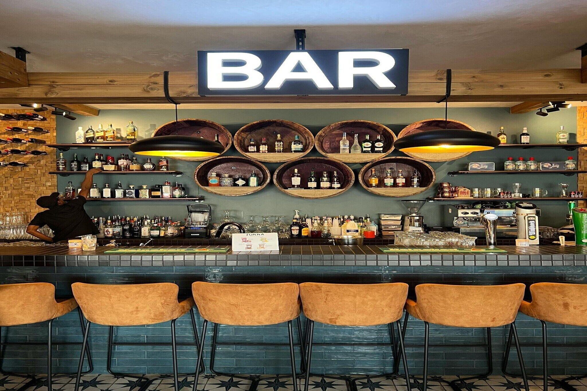 Een bar met een 'BAR' bord, houten planken met flessen en een barman.