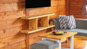 A wooden cabin interior with a flat-screen TV mounted on the wall, a wooden table with a bench, and a sofa with patterned cushions. - Diamond Cabin House (Dieng Kulon)