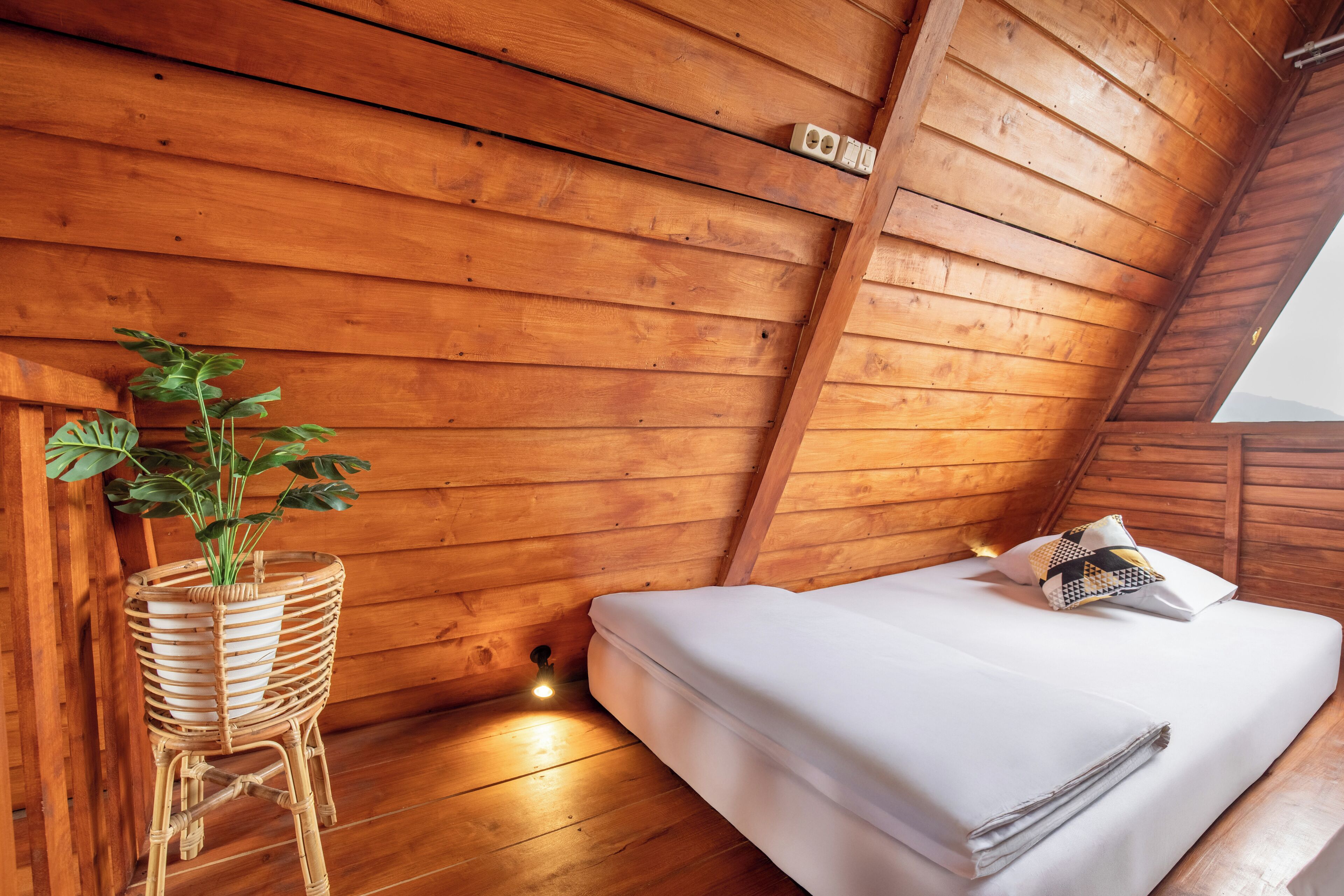 A wooden cabin room with a bed, a wicker plant stand, and a small wall-mounted light.