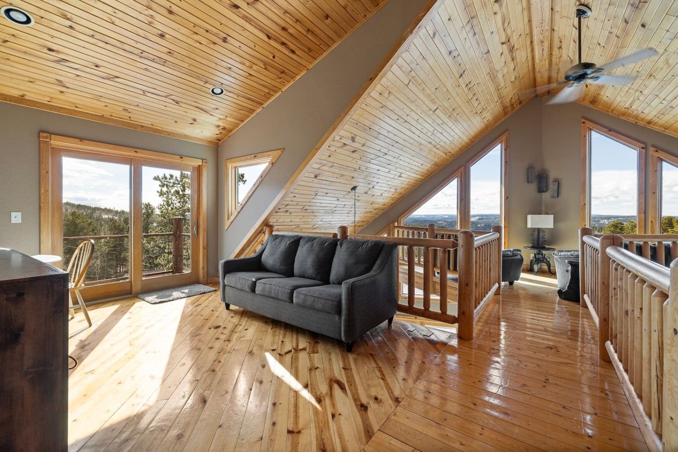 A spacious loft with wooden floors, a grey sofa, and large windows offering a view of the outdoors.