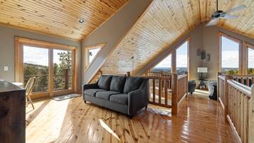 A spacious loft with wooden floors, a grey sofa, and large windows offering a view of the outdoors.