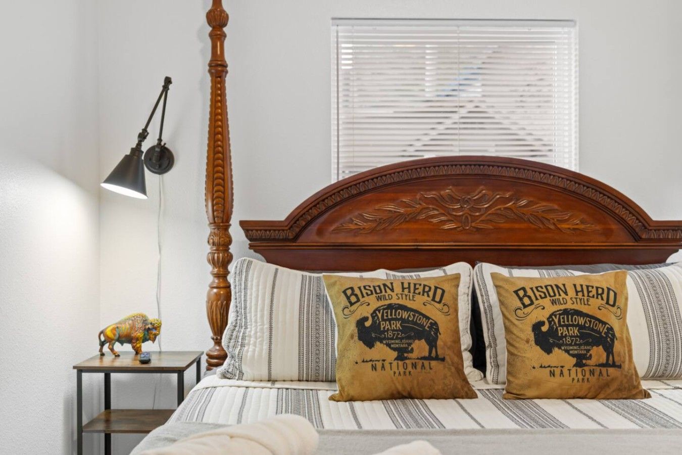 Une chambre à coucher avec une tête de lit en bois, deux oreillers ornés de la mention « Bison Herd », une lampe et une petite table sur laquelle est posée une figurine.