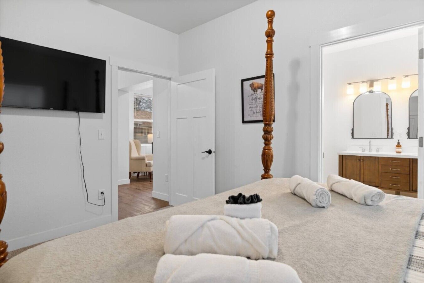 A bedroom with a large bed, a television mounted on the wall, a wooden chair, and a bathroom with a mirror and sink.