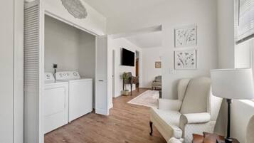 Ein modernes Wohnzimmer mit einer Couch, einem Couchtisch und einem Fernseher. Es gibt eine Waschküche mit Waschmaschine und Trockner. Der Raum hat Holzdielenböden und weiße Wände.