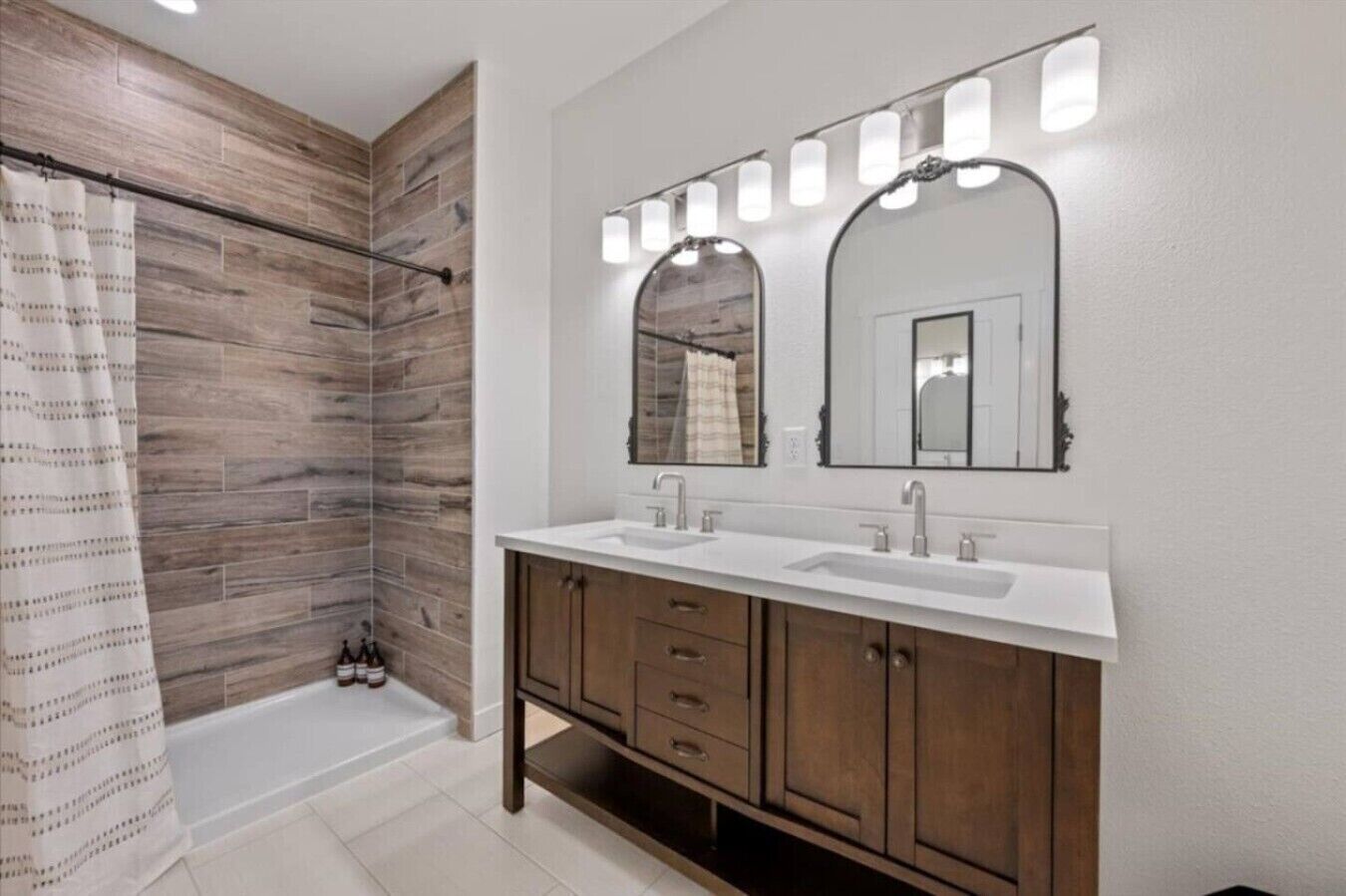 A bathroom with a shower, two sinks, wooden cabinets, and a mirror with lights.