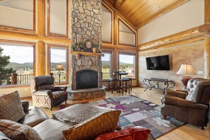 A spacious living room with a stone fireplace, leather furniture, and large windows offering a view of the outdoors. - Golf Course, Sleeps 12, Garage (Sturgis)