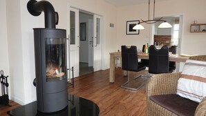 A modern living room with a wood-burning stove, a dining area with a wooden table and chairs, and a wicker sofa.
