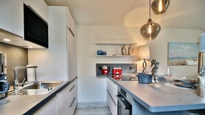 Une cuisine moderne équipée d’un four à micro-ondes encastré, d’une machine à café et d’un évier. On y trouve également un coin repas avec une table pour deux personnes, un luminaire suspendu et une étagère avec des verres et une bouilloire.