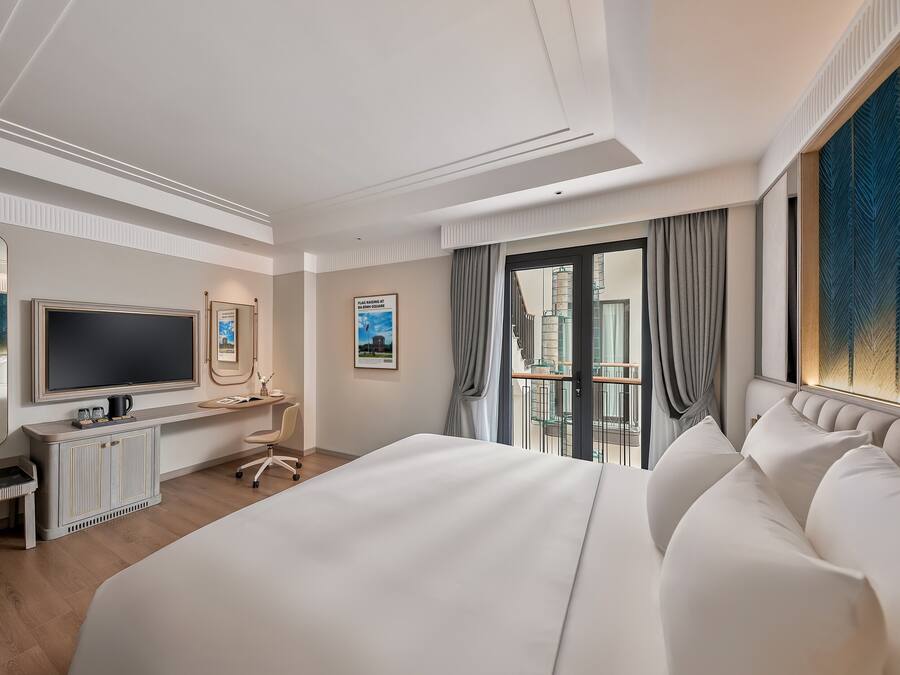 A modern hotel room with a large bed, a desk with a flat-screen TV, a seating area, and a balcony with a city view.