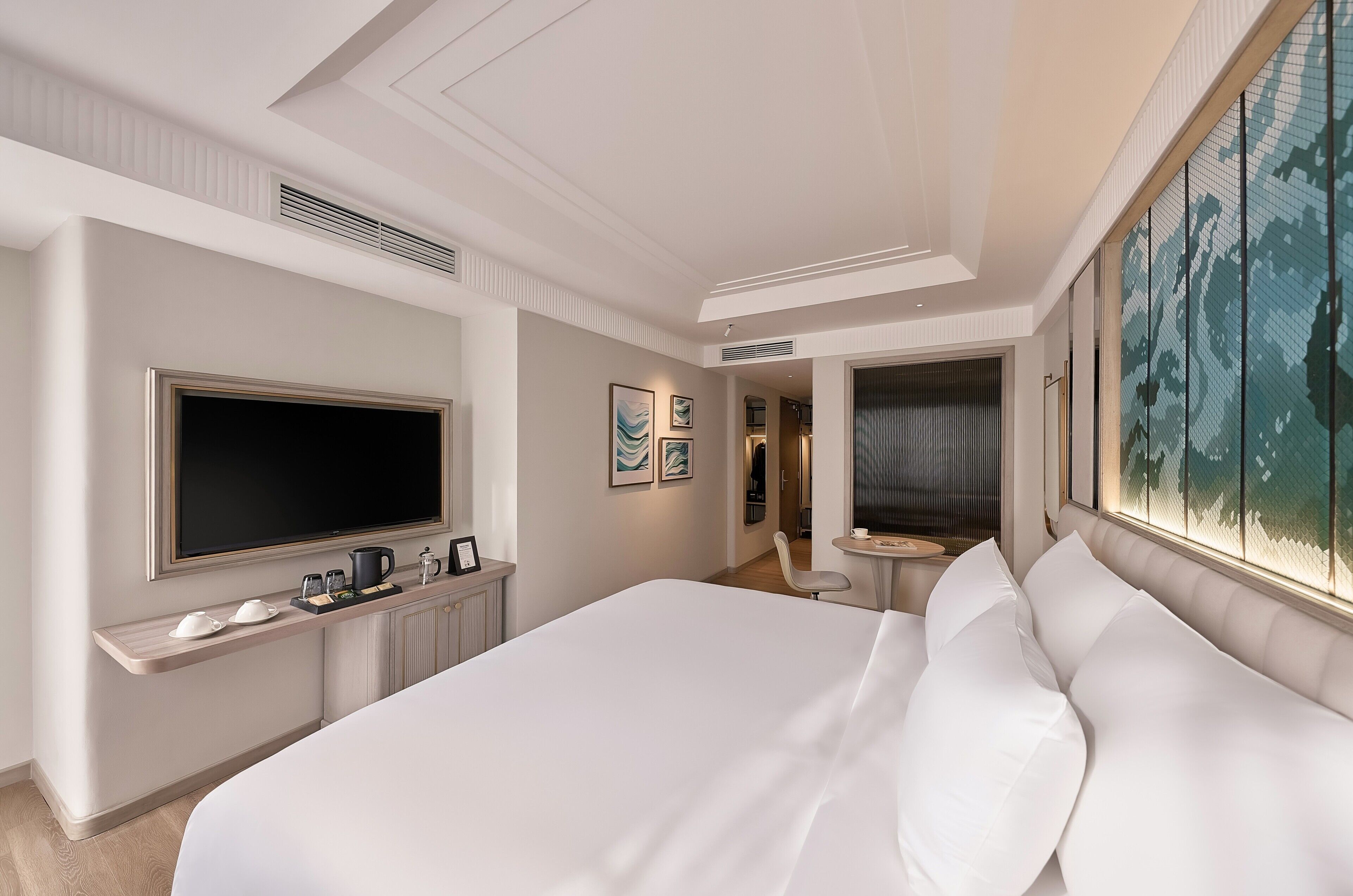 Habitación de hotel con una cama grande, un televisor de pantalla plana, una mesita pequeña con una tetera y tazas, y obras de arte en las paredes.