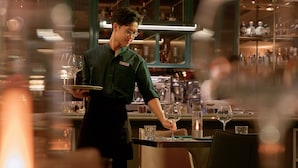 Een restaurantinterieur met een ober die een klant bedient. Er zijn tafels, stoelen en een bar te zien.