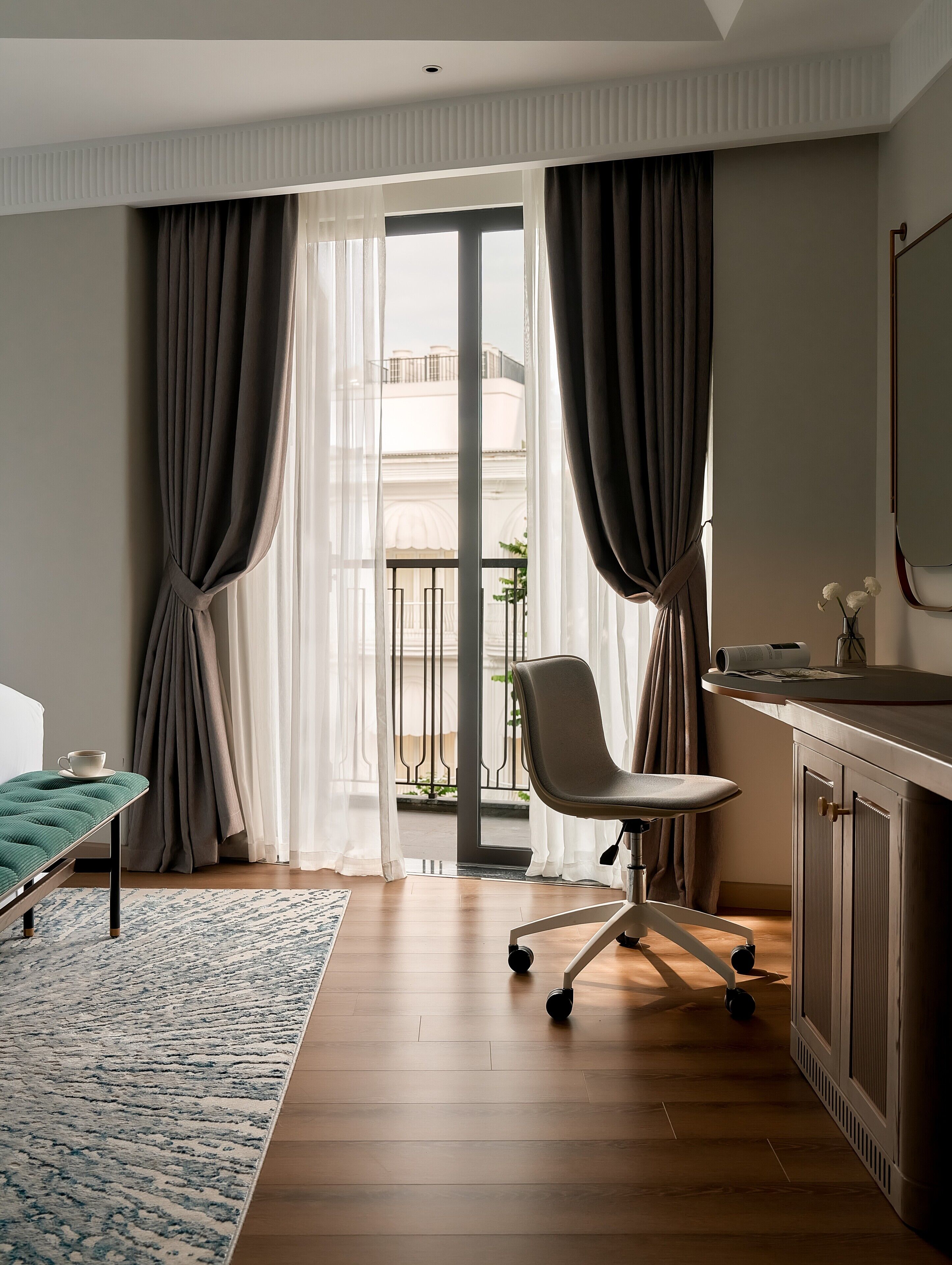 A hotel room with a bed, a desk, a chair, a small table, and a view of a building through sheer curtains.