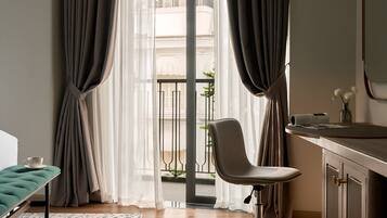 Habitación de hotel con cama, escritorio, silla, una mesita y vistas a un edificio a través de cortinas translúcidas.