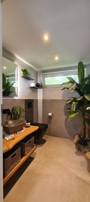 A modern bathroom with a large mirror, a stone basin, and a black toilet. There are plants and a window providing natural light. - Modern vacation bungalow in the Neuhof forest settlement on the Großer Wünsdorfer See lake (Zossen (Neuhof))