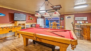 Una mesa de billar con superficie de fieltro rojo, una mesa de fútbol de mesa y una zona de bar con un refrigerador y una televisión.
