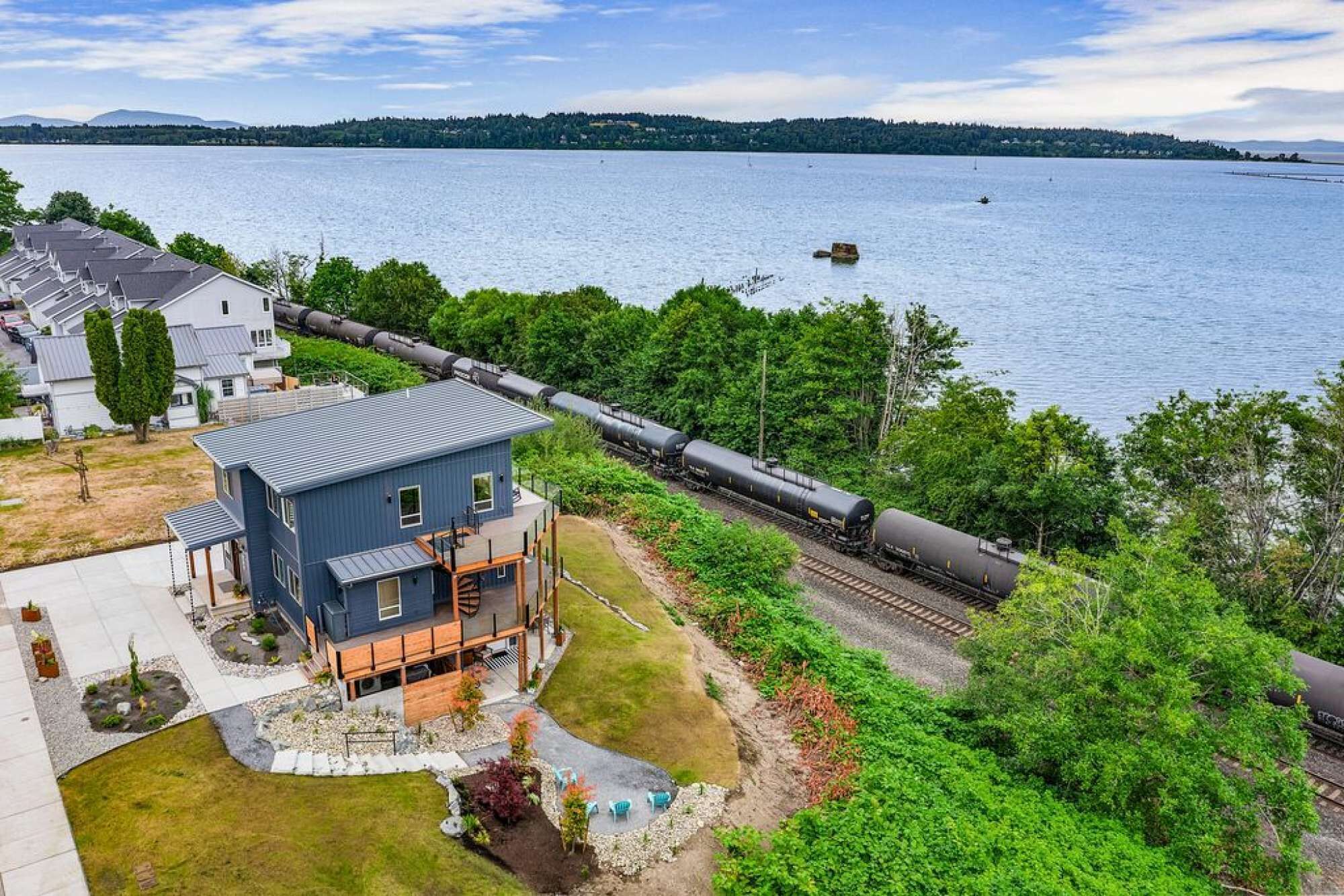 A modern two-story house with a balcony, located near a waterfront with a train passing by.