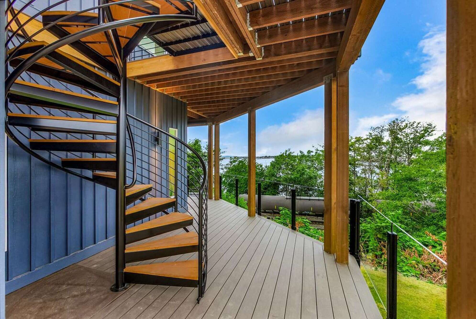 A spiral staircase with wooden steps and metal railings, leading to a covered outdoor area with a wooden deck and a view of greenery and a body of water.