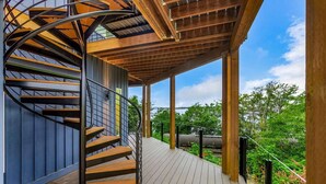 Un escalier en colimaçon avec des marches en bois et des rampes en métal, menant à un espace extérieur couvert doté d’une terrasse en bois et offrant une vue sur la verdure et un plan d’eau.