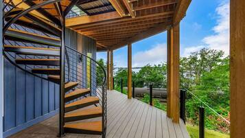A spiral staircase with wooden steps and metal railings, leading to a covered outdoor area with a wooden deck and a view of greenery and a body of water.
