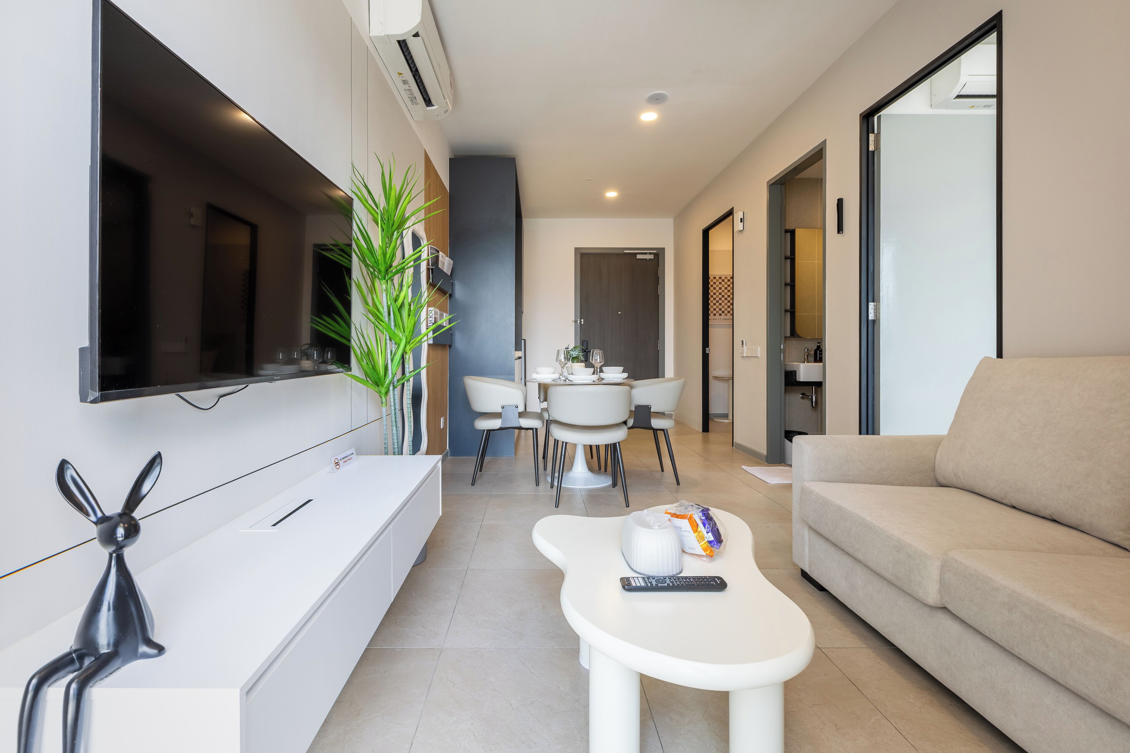 Una sala moderna con un sofá, una mesa de centro blanca con un control remoto y bocadillos, un televisor montado en la pared y un comedor con mesa y sillas.
