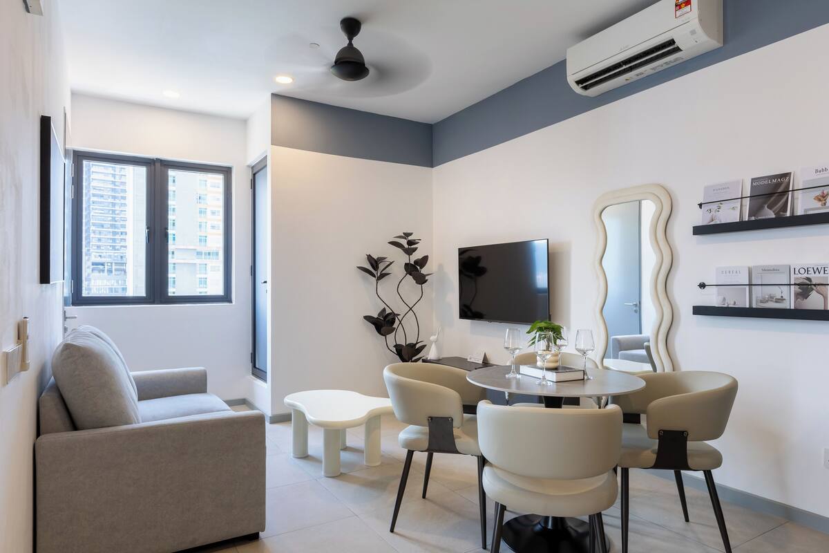 A modern living room with a sofa, dining table, and chairs. There is a flat-screen TV mounted on the wall, a ceiling fan, and an air conditioning unit.