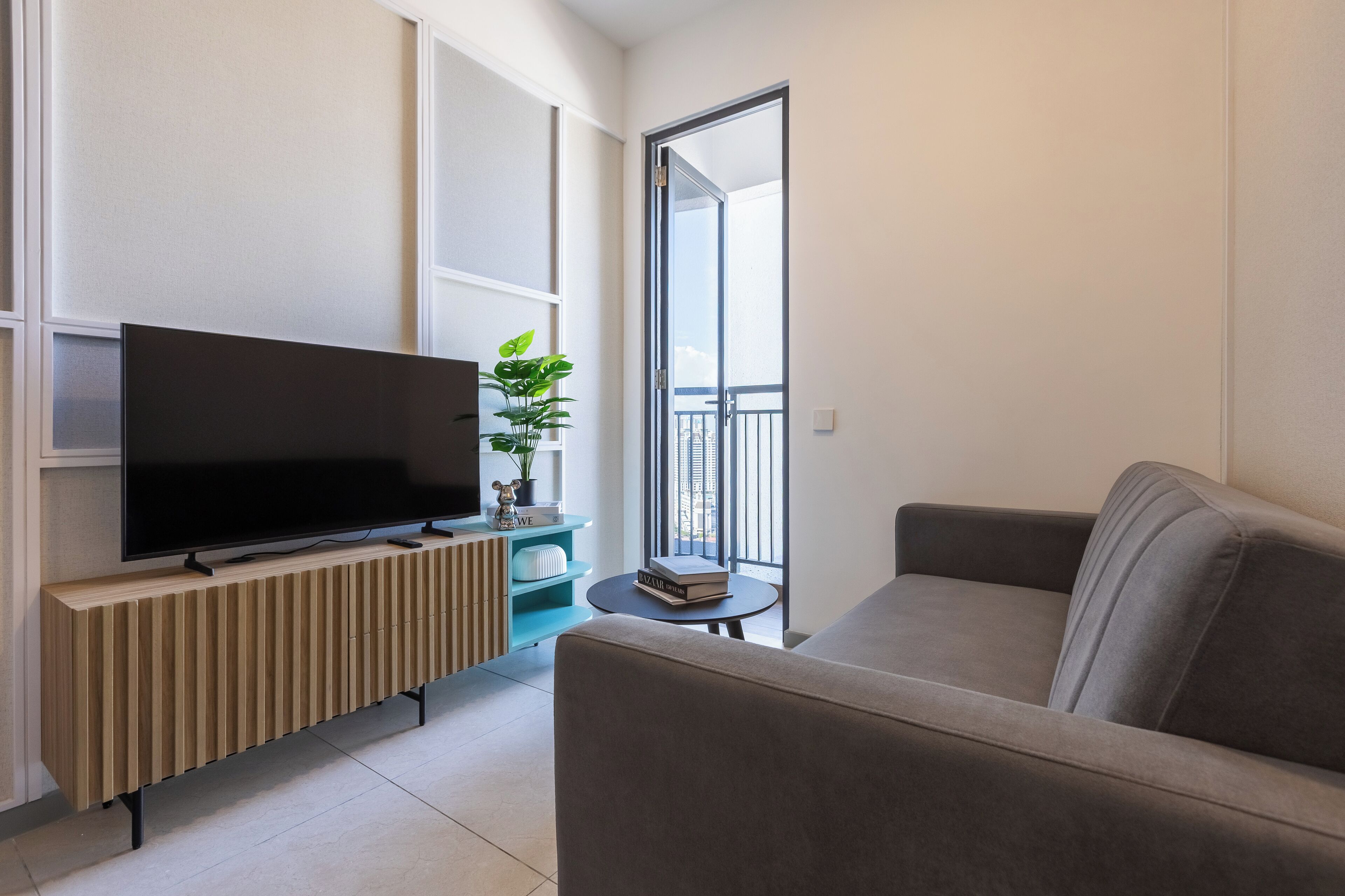 Una sala moderna con un televisor de pantalla plana sobre un mueble de madera, un sofá gris, una mesita redonda con una planta y un balcón con barandilla.