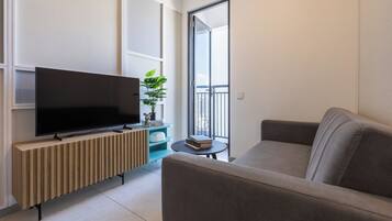 Una sala moderna con un televisor de pantalla plana sobre un mueble de madera, un sofá gris, una mesita redonda con una planta y un balcón con barandilla.