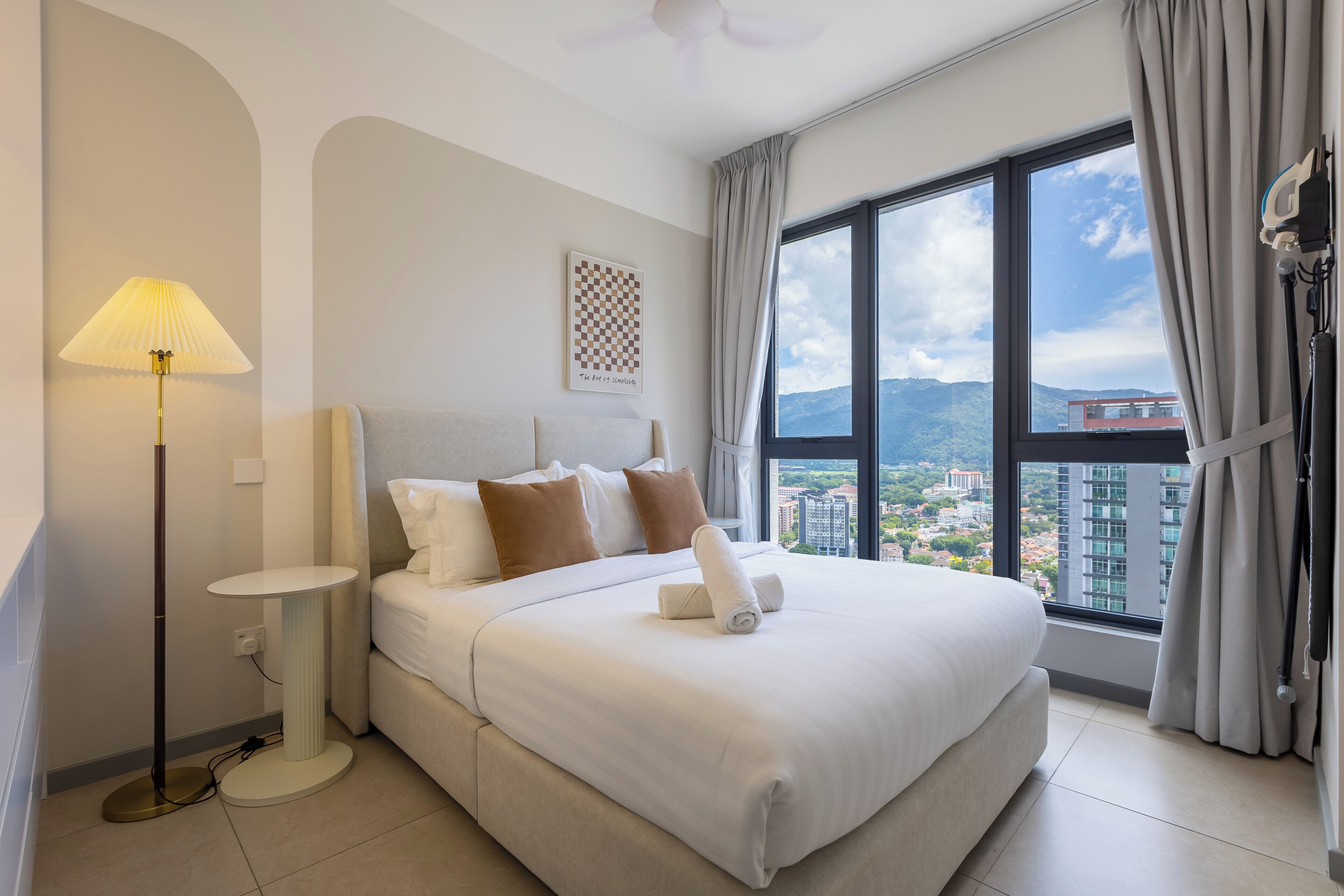 A hotel room with a large bed, a bedside table, a lamp, a window with curtains, and a view of the city.