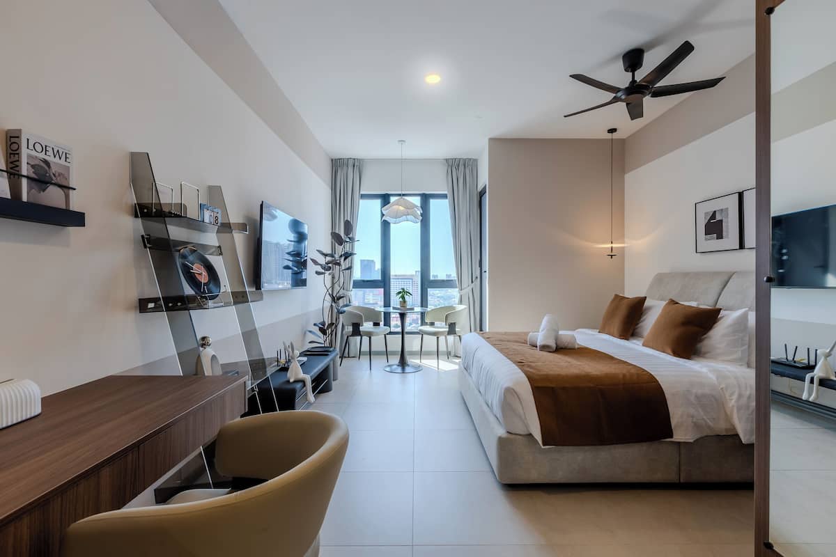 A modern hotel room with a bed, a desk, a chair, a small table, and a large window with a city view.