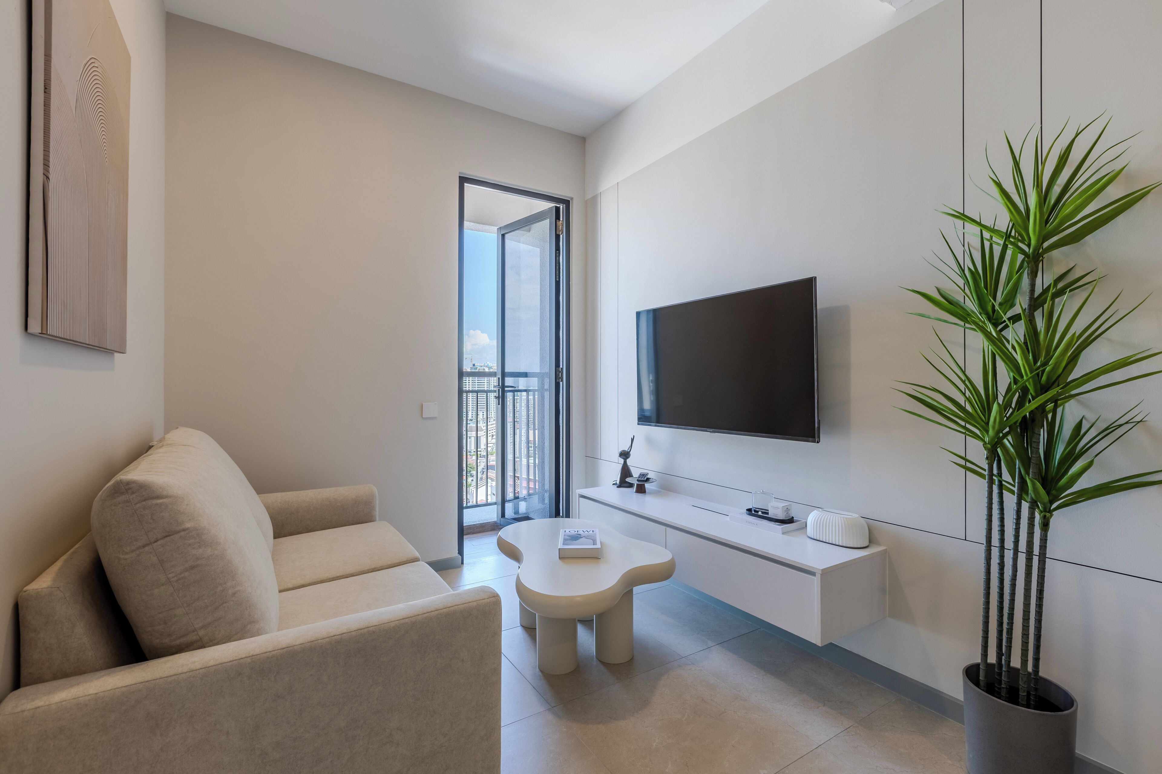 A modern living room with a sofa, a coffee table, a flat-screen TV, a potted plant, and a view of the outdoors through a glass door.