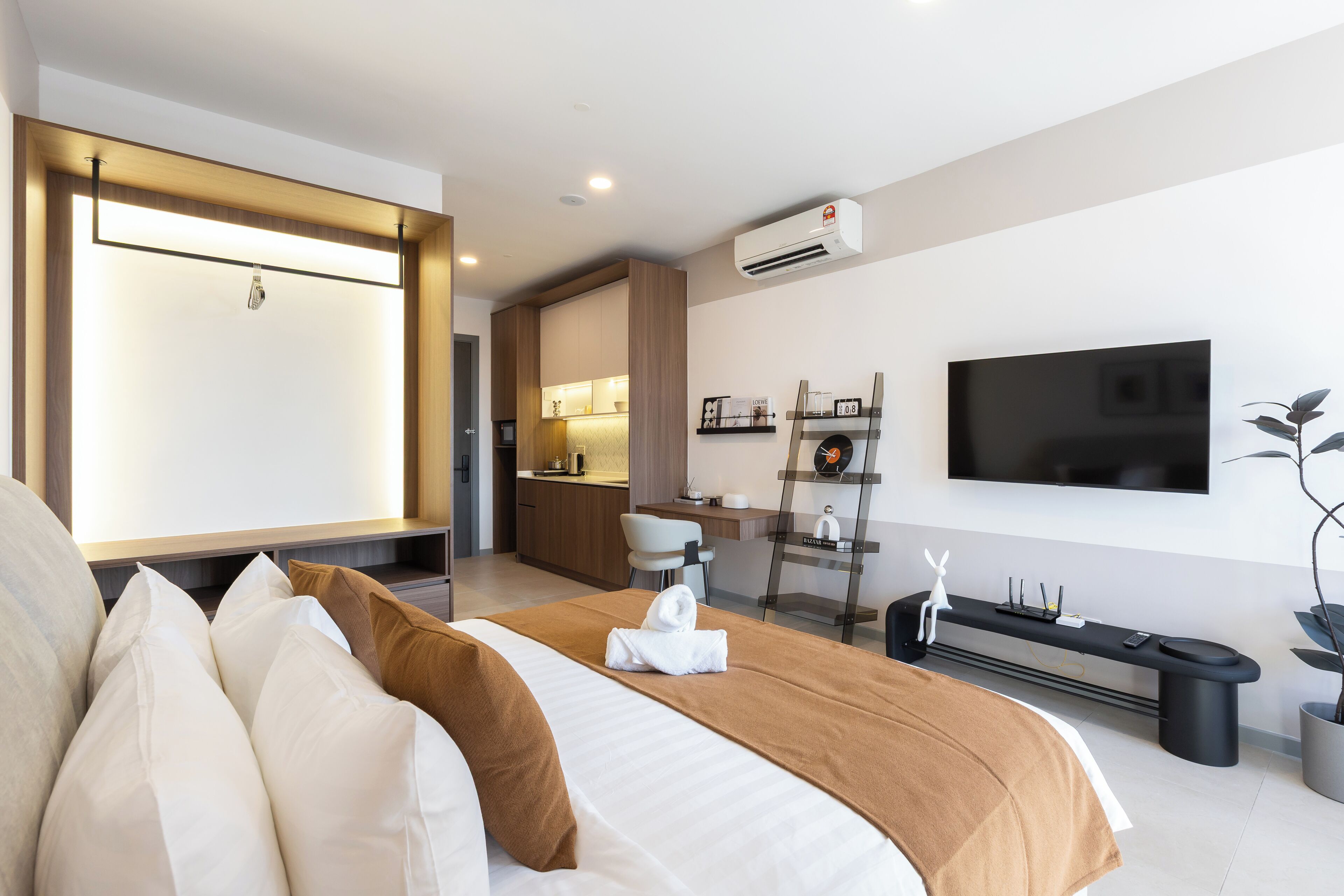 A modern hotel room with a large bed, a flat-screen TV, a desk with a chair, a small kitchen area, and a potted plant.