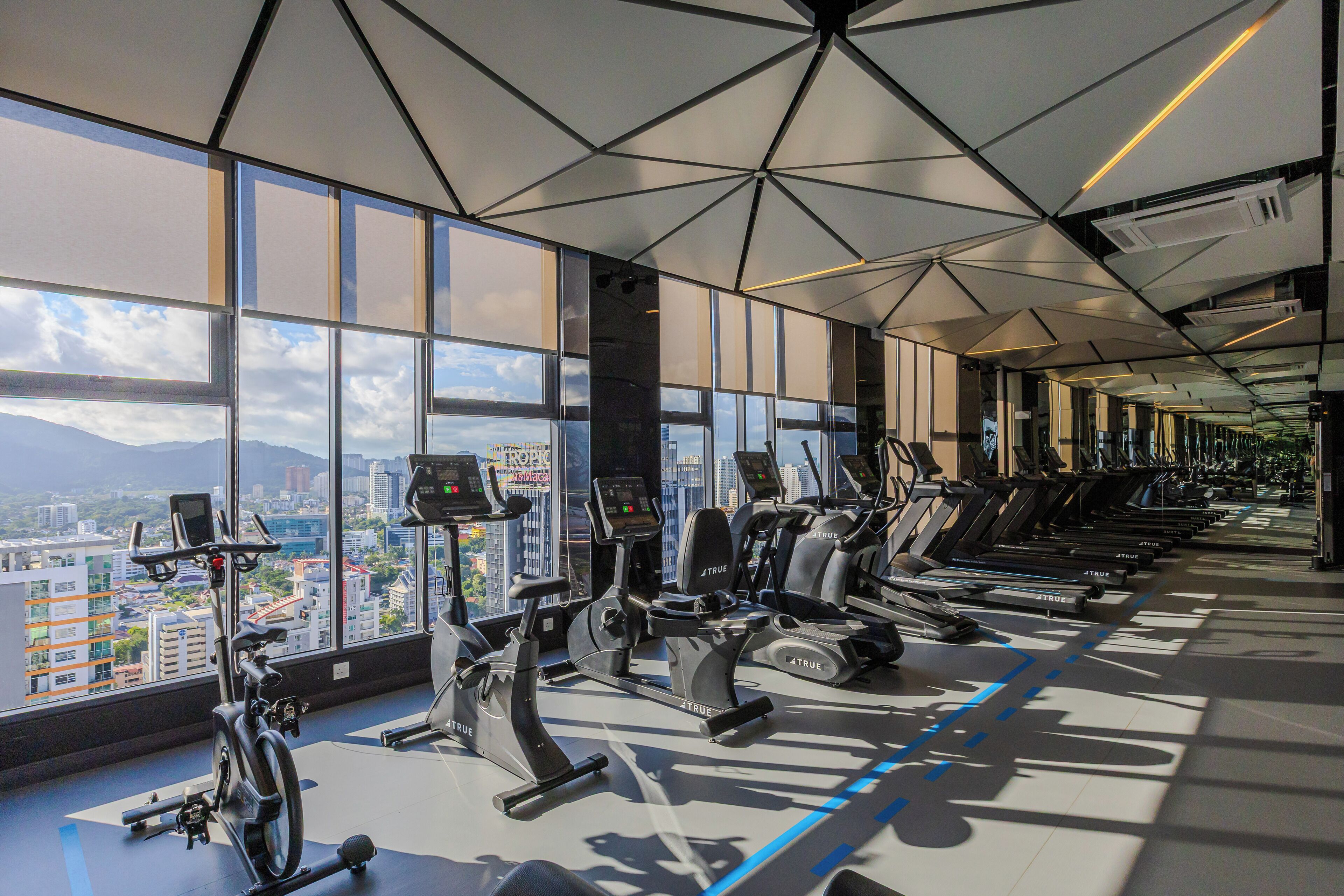 Une salle de sport équipée de divers appareils d’exercice, dotée de grandes fenêtres et offrant une vue sur la ville.