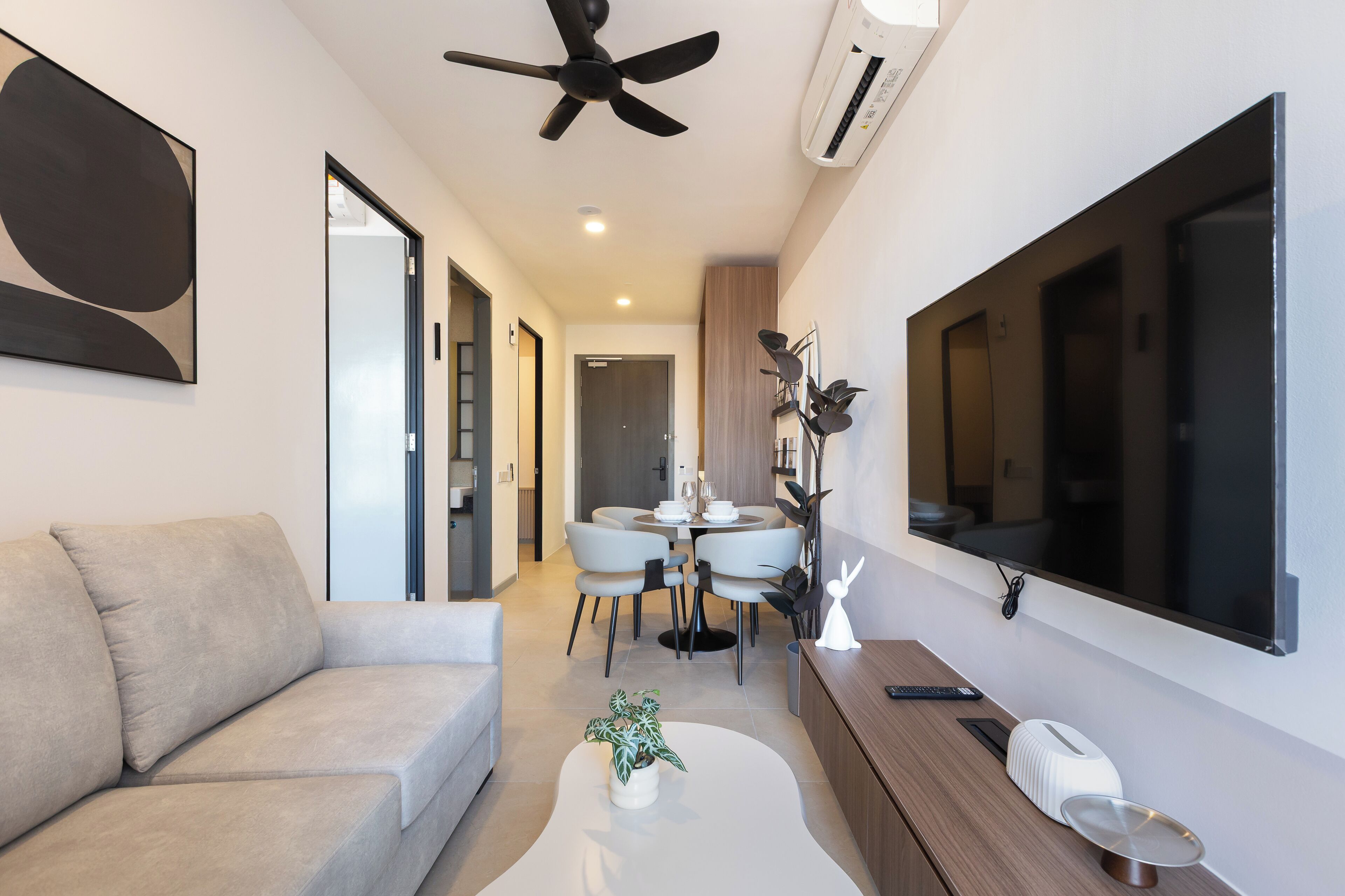 A modern living room with a sofa, a coffee table with a plant, a dining area with a table and chairs, and a large TV mounted on the wall.