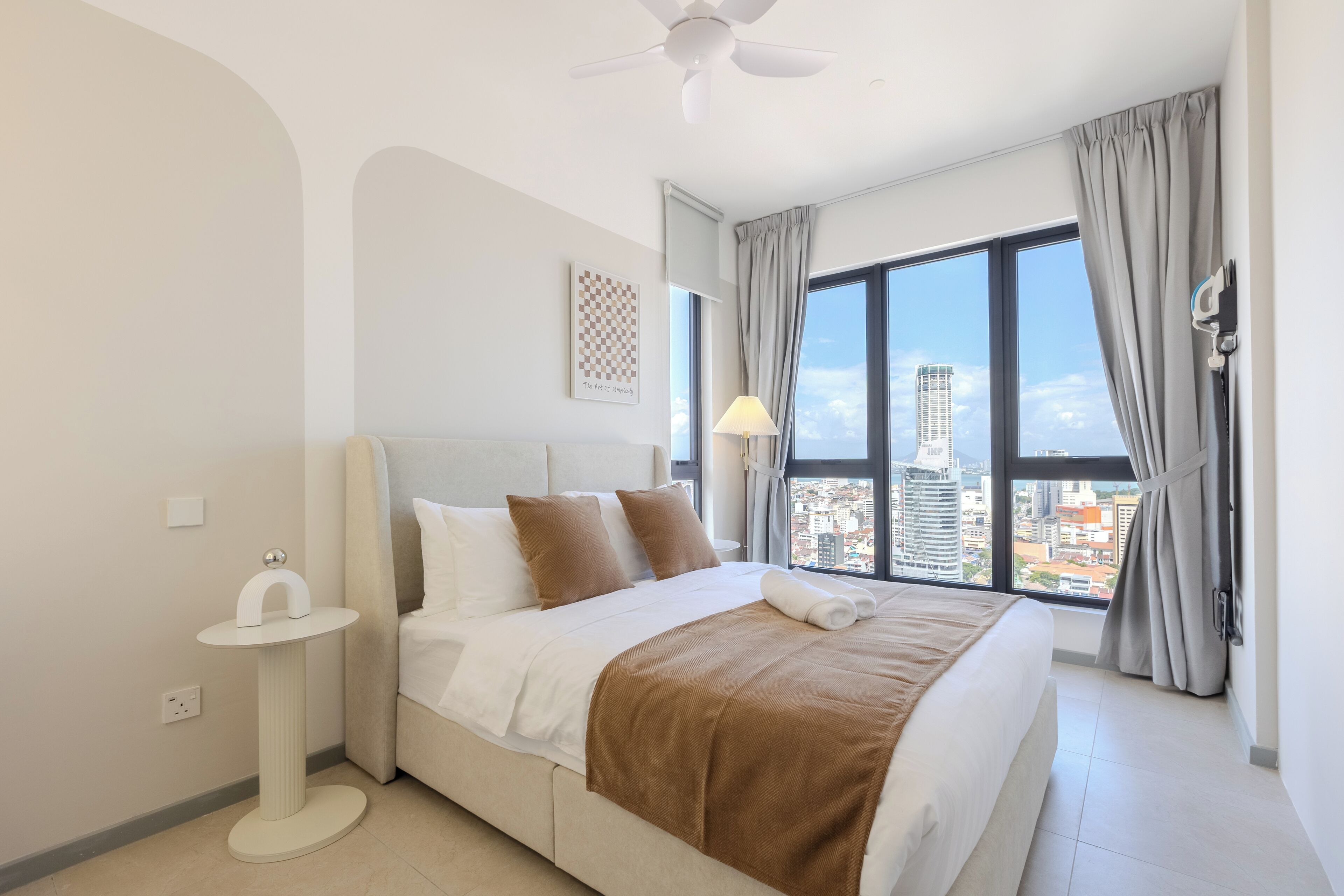 A modern hotel room with a large bed, a bedside table, a ceiling fan, and a city view through large windows.