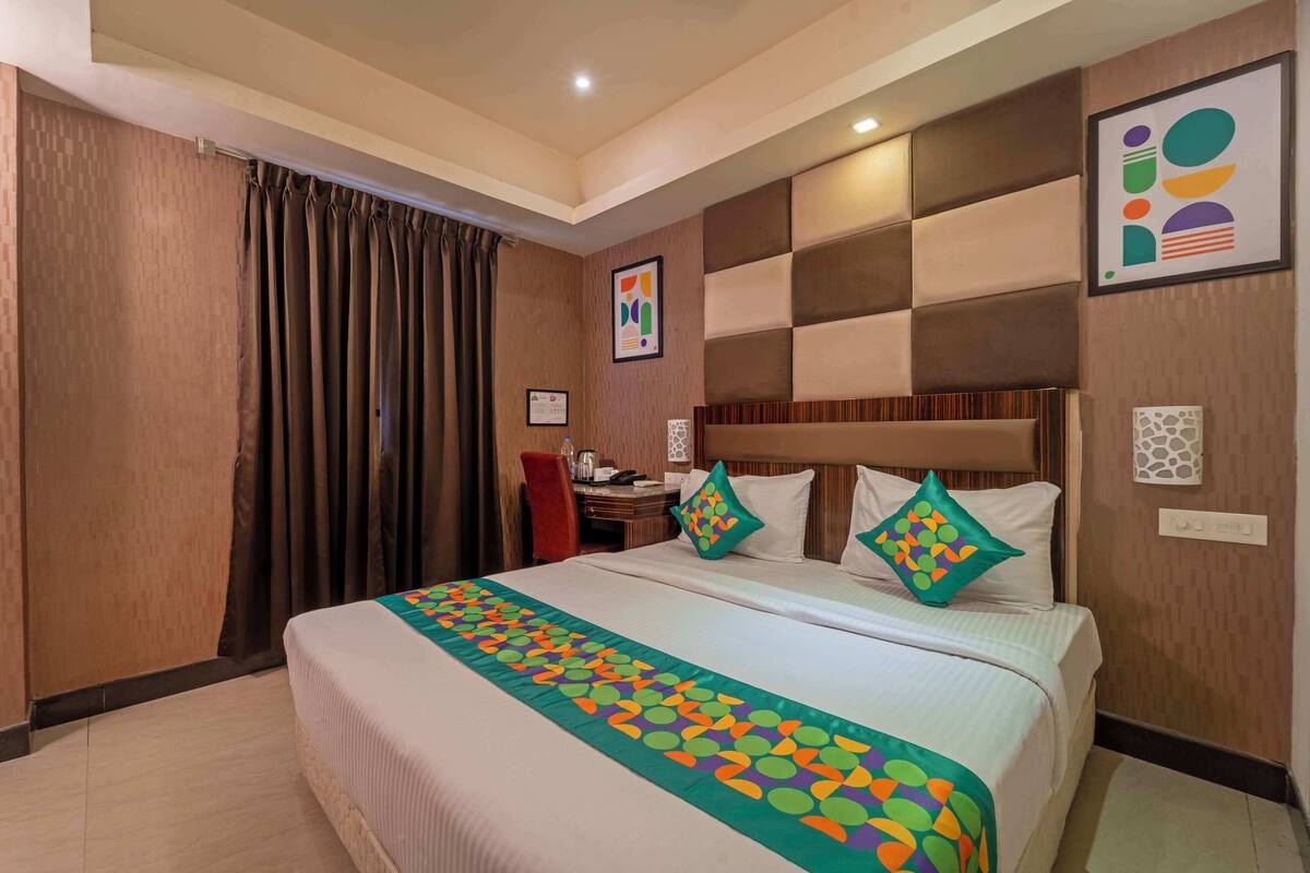 A hotel room with a large bed, a desk, and a chair. There are curtains on the window and a framed picture on the wall.