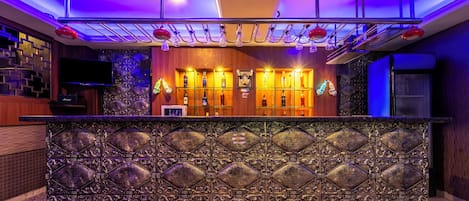 Una zona de bar con frente metálico decorativo, estantes con botellas y un televisor.