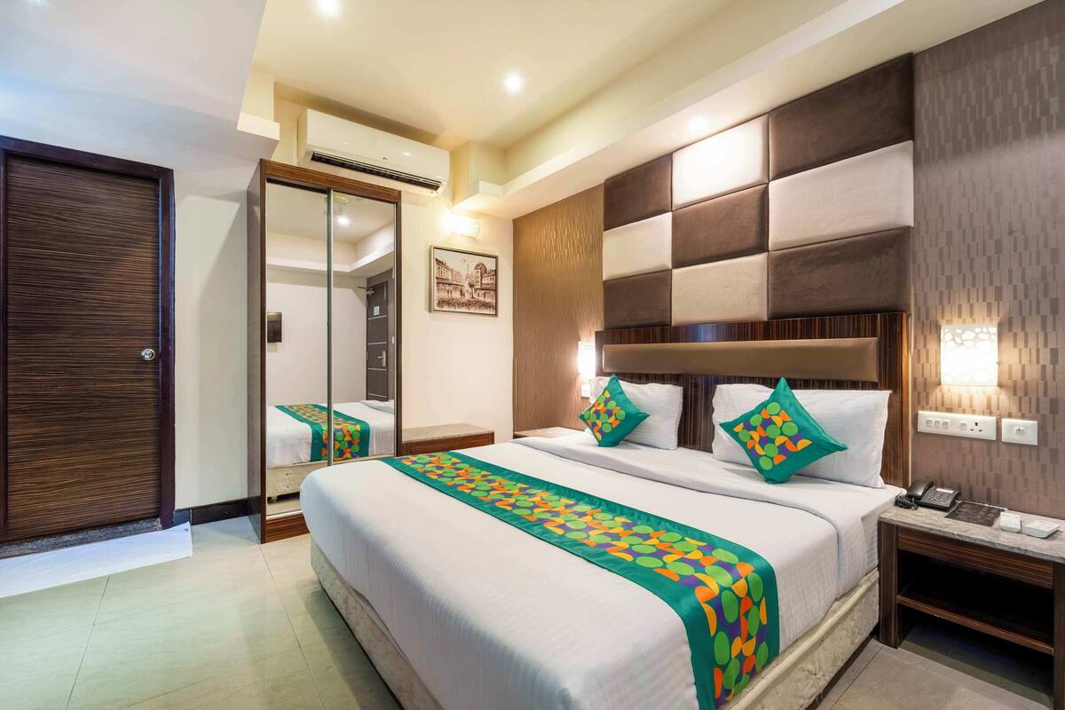 A hotel room with a large bed, a bedside table with a phone and a lamp, a wardrobe, and a mirror.