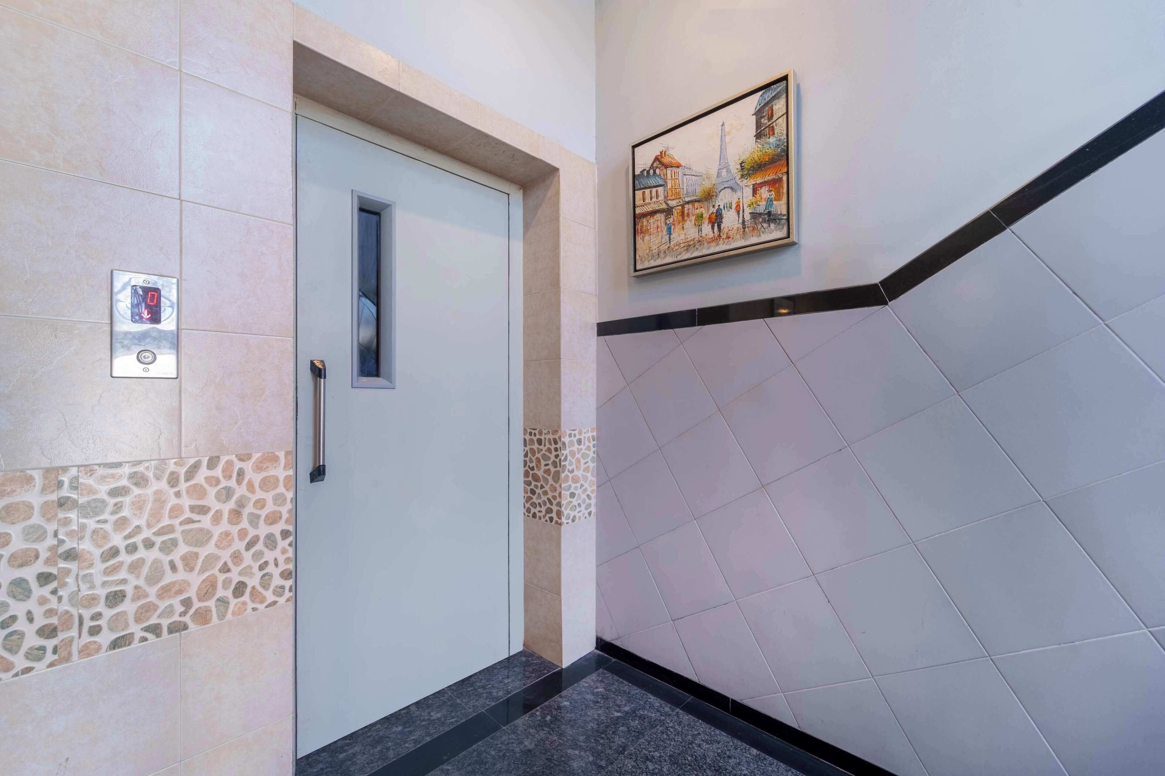 Un pasillo con una puerta, un panel de control de ascensor en la pared, una sección de suelo de baldosas estampadas y un cuadro con una vista urbana.